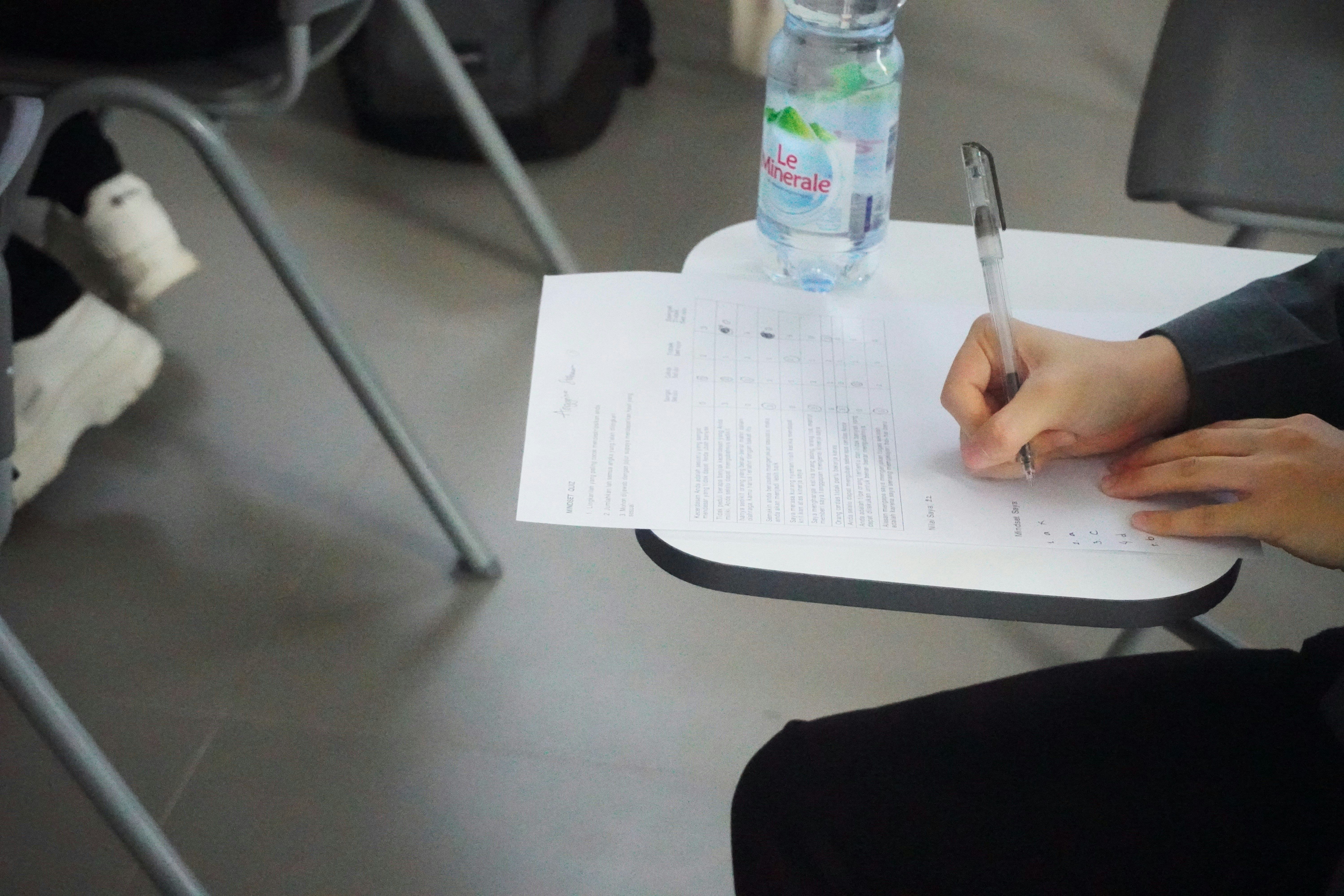 A man attempting an exam in a classroom on a piece of paper using a ball point pen with a water bottle next to her.