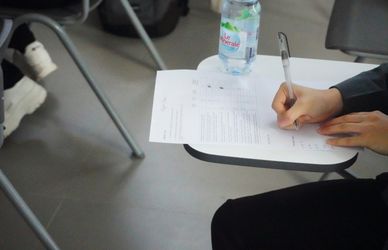 A man attempting an exam in a classroom on a piece of paper using a ball point pen with a water bottle next to her.